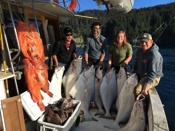Four people pose on a boat with a large catch of bright orange rockfish and several large white halibut.