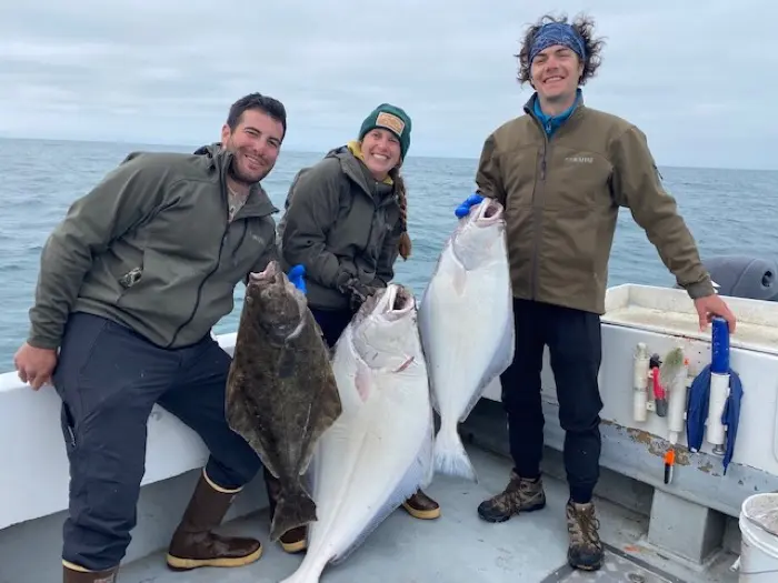 Three people on a boat deck smile while holding three large, flat fish, likely halibut, caught at sea.