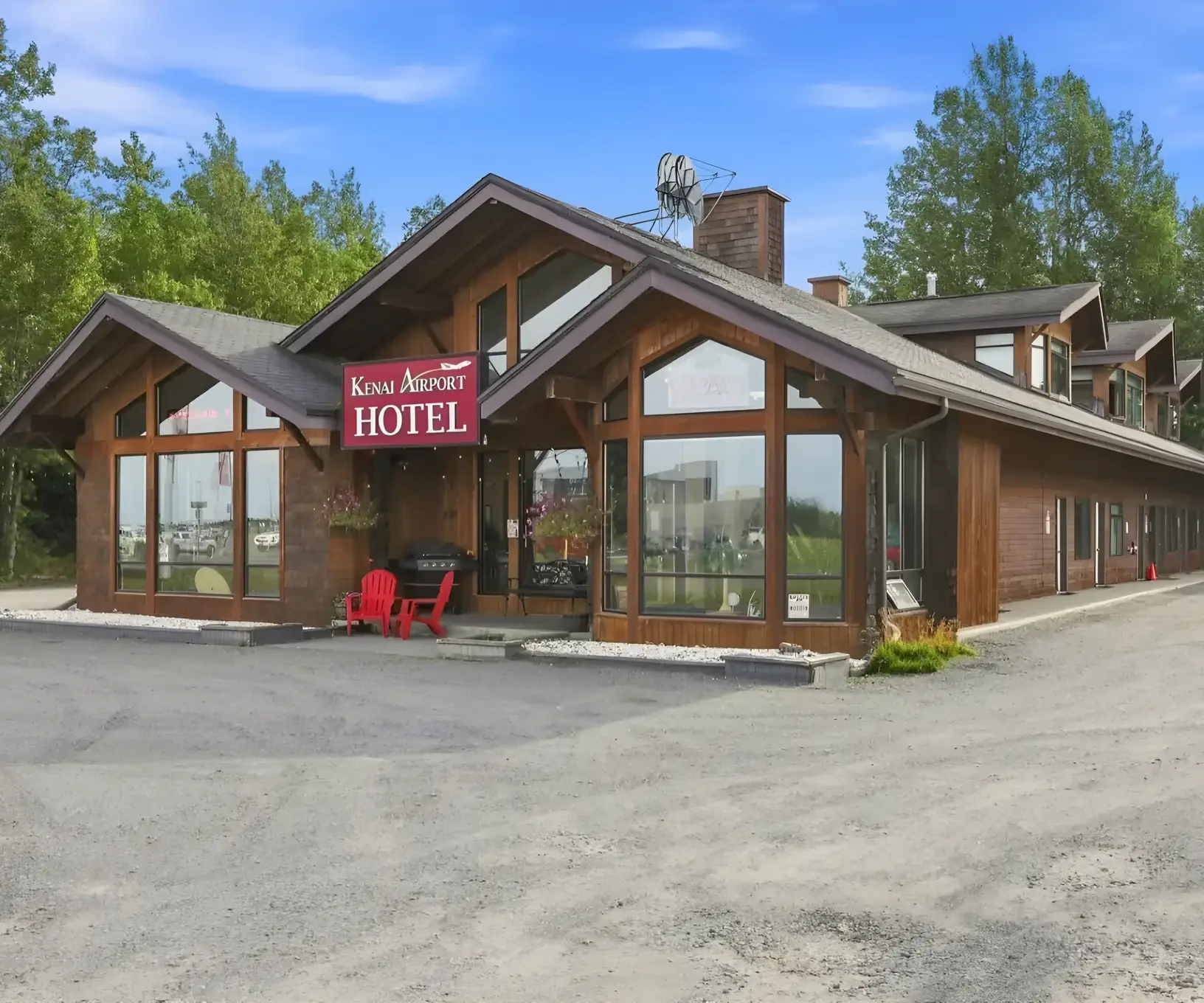 The Kenai Airport Hotel is a wooden, lodge-style building with large windows, surrounded by trees on a gravel lot.