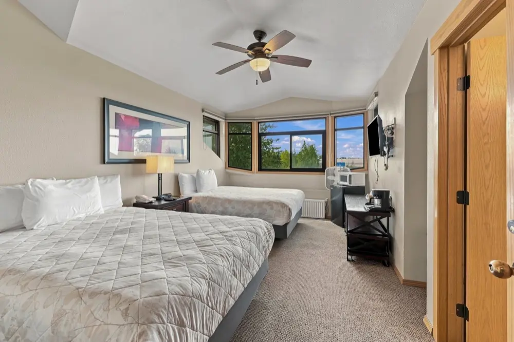 A hotel room features two beds with white quilted bedding, a ceiling fan, a window, and a door on the right.