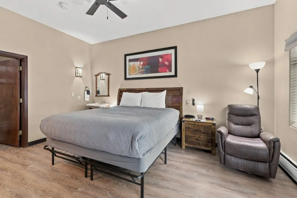 A hotel room with a queen bed, a wood nightstand, a gray recliner, a floor lamp, and a framed piece of artwork.
